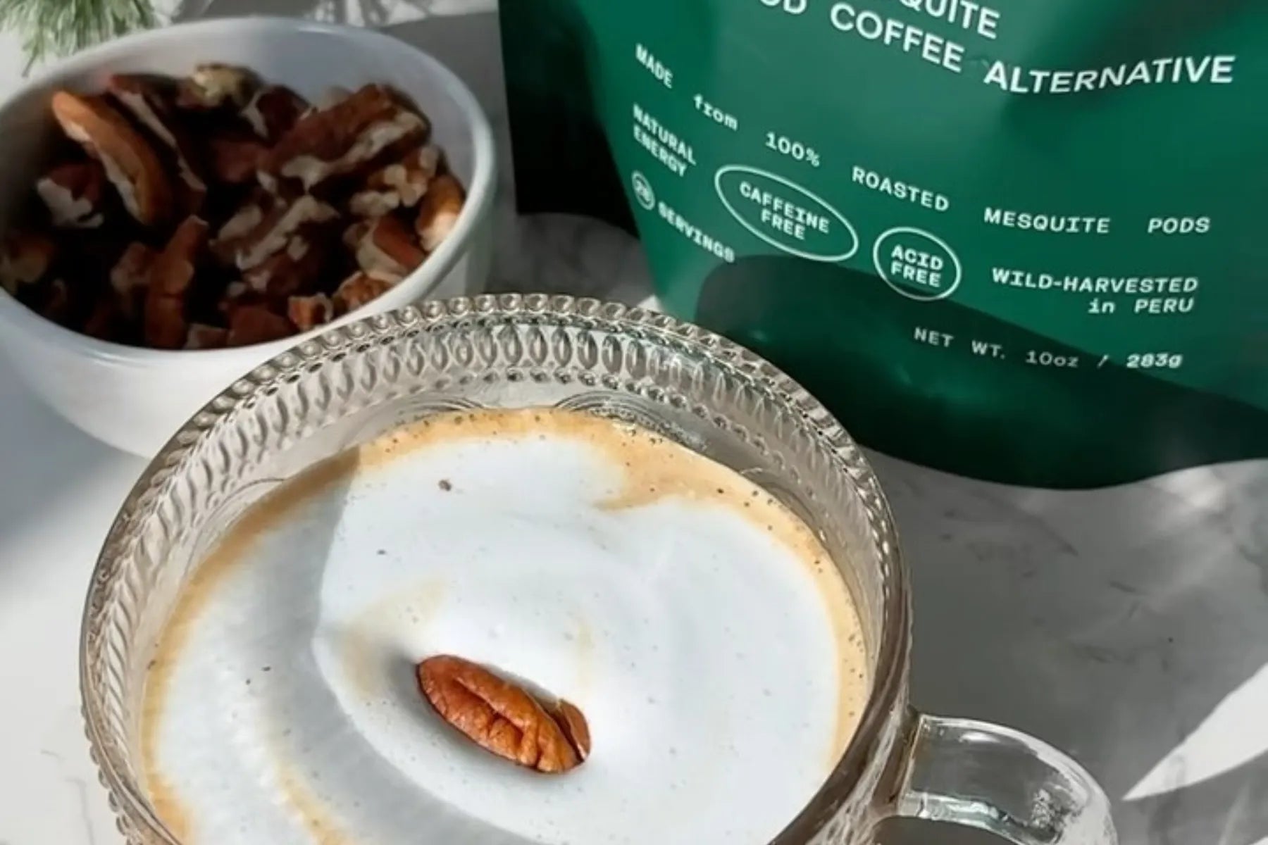 Coffee alternative drink in a glass mug topped with foam and a pecan, alongside a bowl of nuts and packaging.