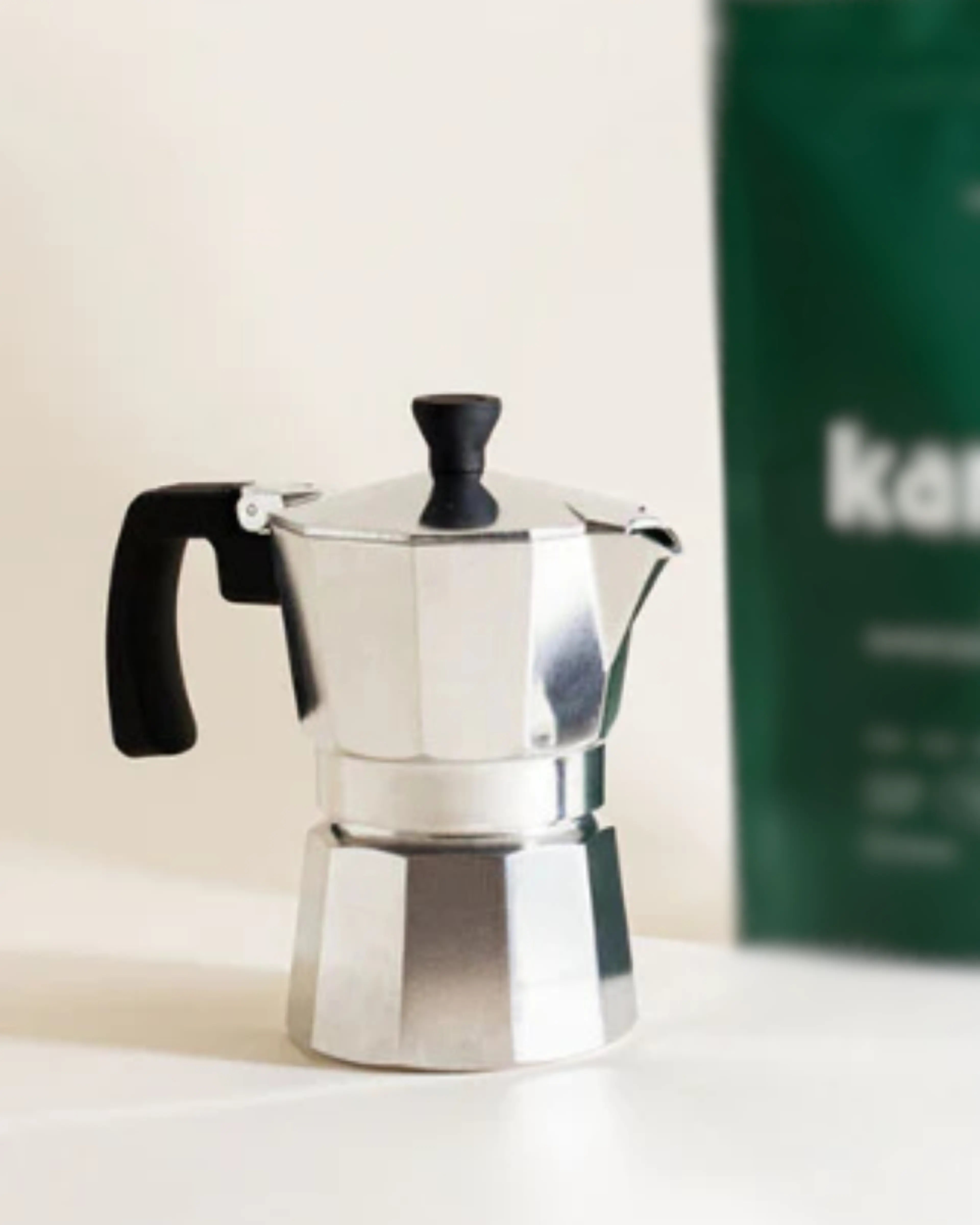 Moka pot with a black handle on a white surface, Kamana Mesquite Superfood Coffee package in the background.